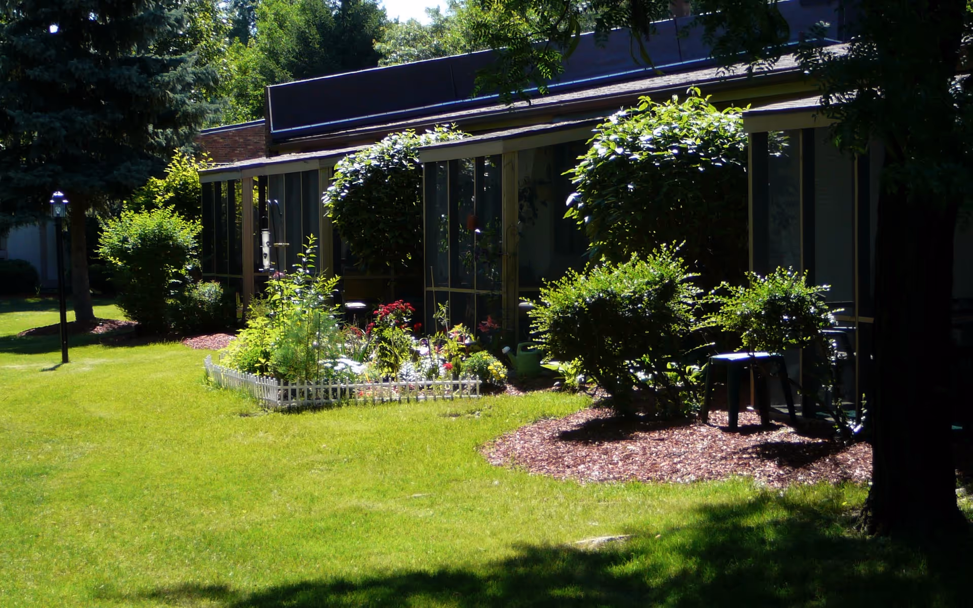 Sunlit landscaped lawn and flower beds in front of a low building with screened porches and shrubs.