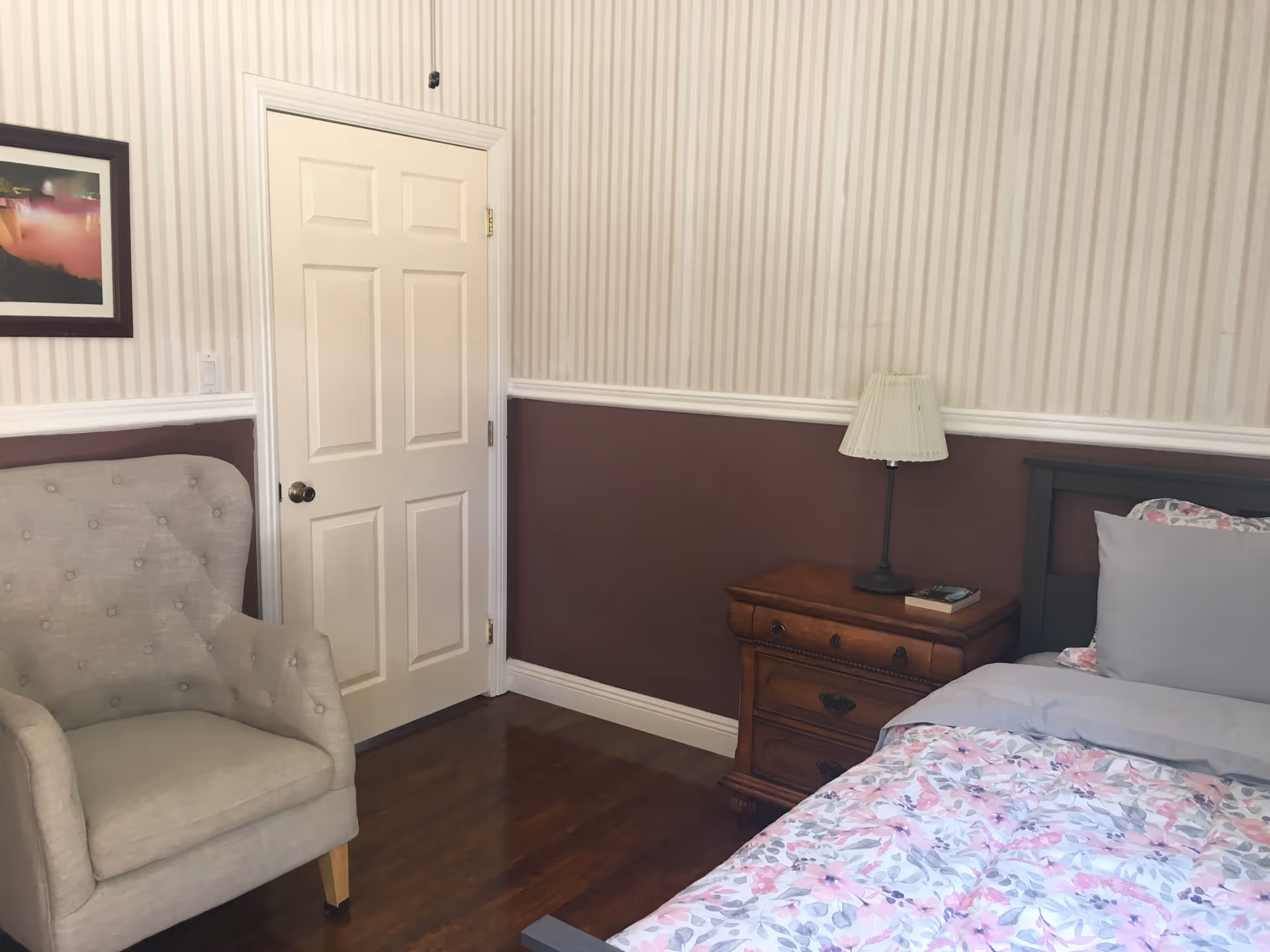 A cozy bedroom corner featuring a bed with floral bedding and gray pillows, a wooden nightstand with a lamp and a book, a beige upholstered armchair, a closed white door, and a framed picture on the wall. The walls have a two-tone design with striped wallpaper on the upper half and solid maroon paint on the lower half, separated by white trim.