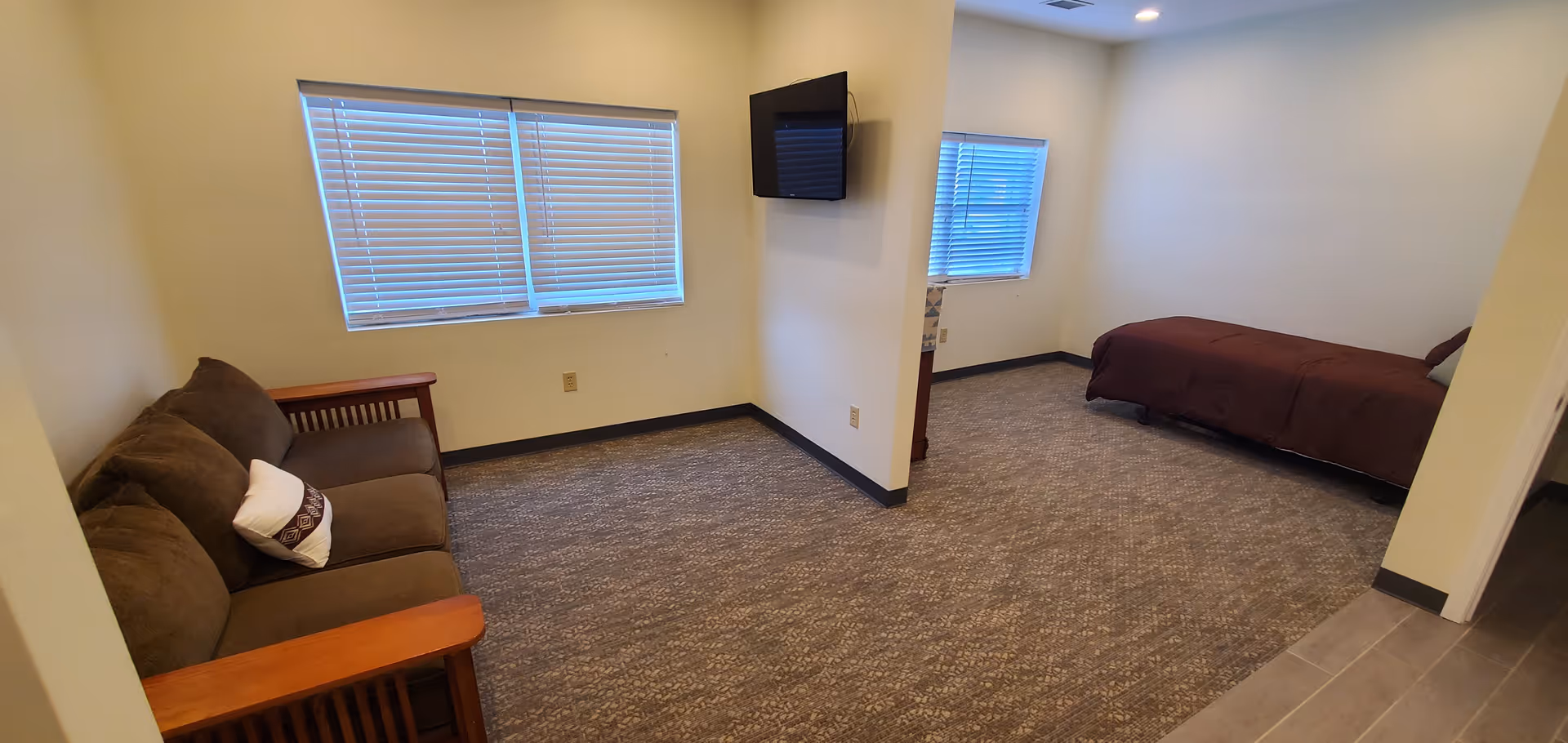 Interior view of a senior living facility room with a brown couch on the left side, a wall-mounted TV in the center, and a single bed with a brown bedspread on the right side. The room has beige walls, carpeted flooring, and windows with closed blinds.