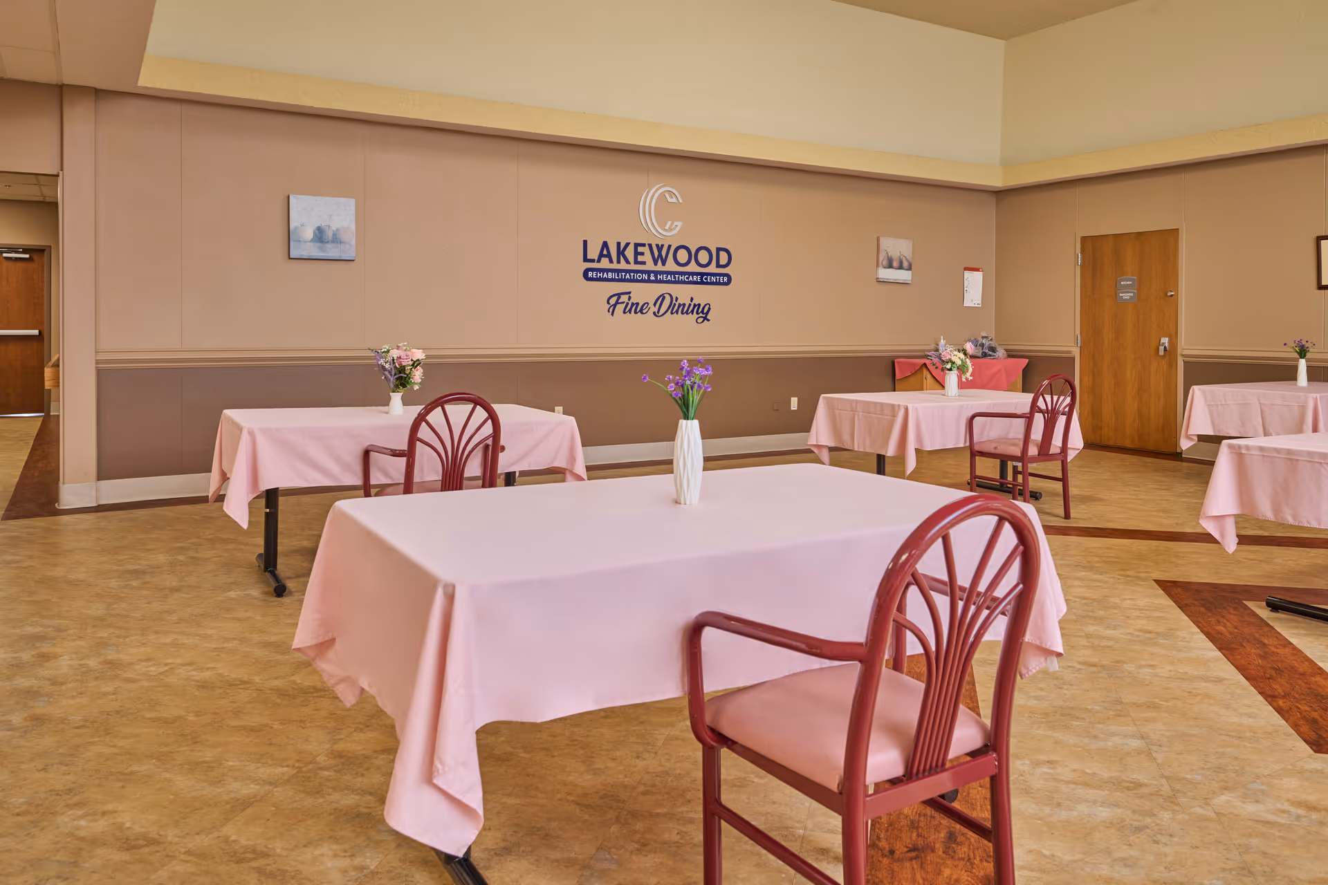Dining room at Lakewood Rehabilitation and Healthcare Center with several tables covered in pink tablecloths, each with a vase of flowers. The walls are beige with the facility's logo and the words 'Fine Dining' displayed prominently. There are red chairs around the tables and a wooden door in the background.