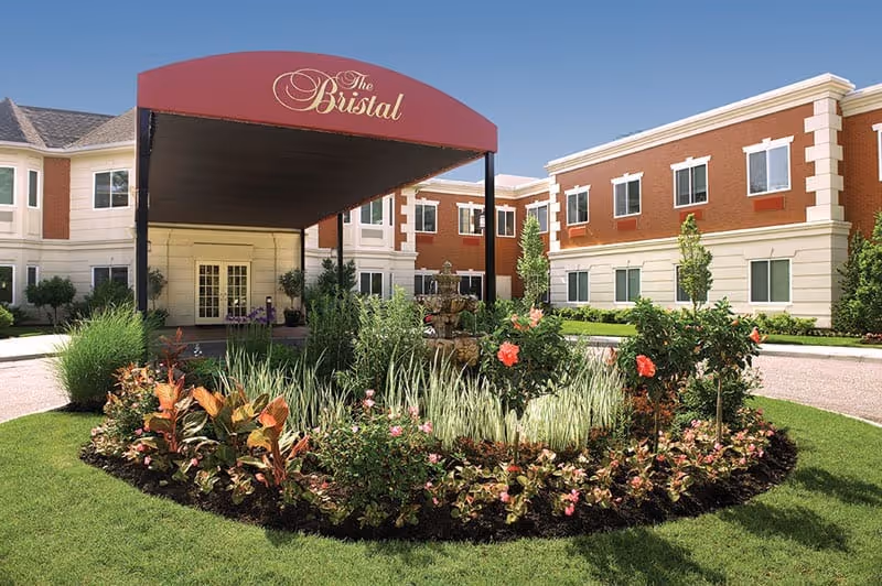 Front exterior of The Bristal Assisted Living at Massapequa with a curved maroon canopy over the entry and a circular landscaped flower bed.