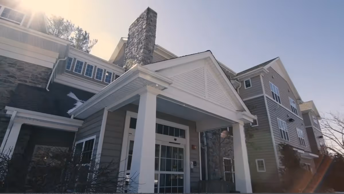 Exterior view of a senior living facility building with a covered entrance supported by white columns, stone chimney, and multiple windows under a clear sky.
