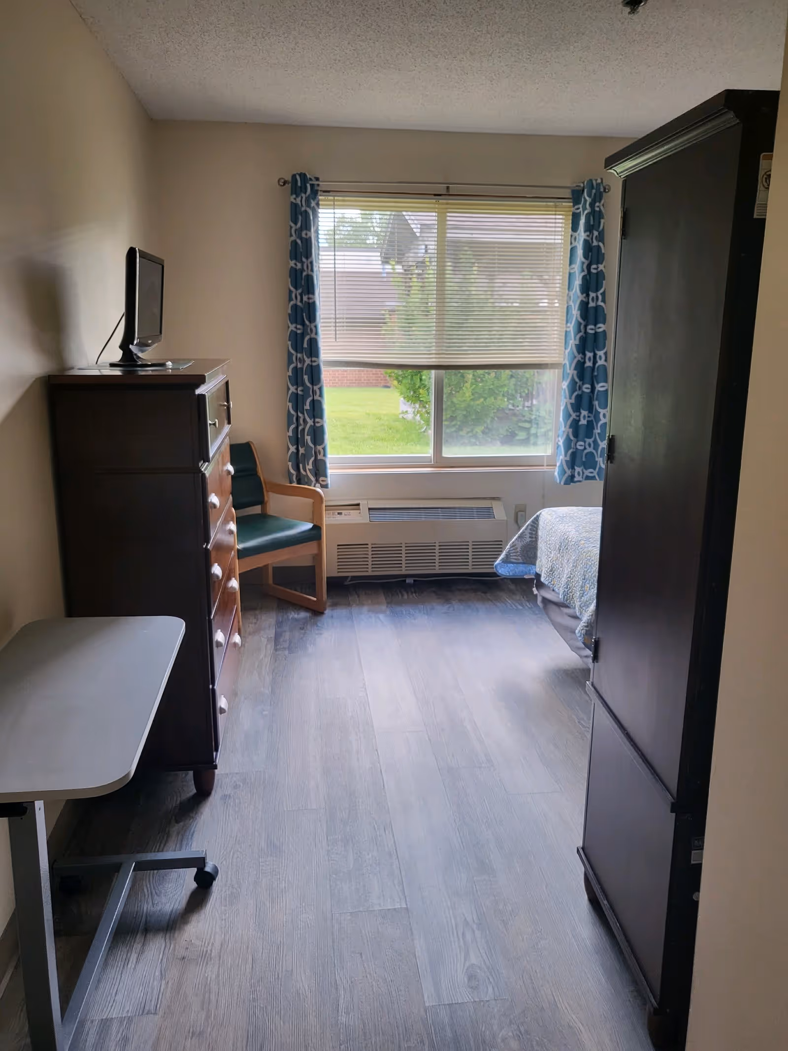 A small bedroom with a wooden dresser topped with a small TV on the left, a green cushioned wooden chair near a window with blue patterned curtains, a heating/cooling unit below the window, and a bed partially visible on the right side. The floor has wood-like vinyl planks.