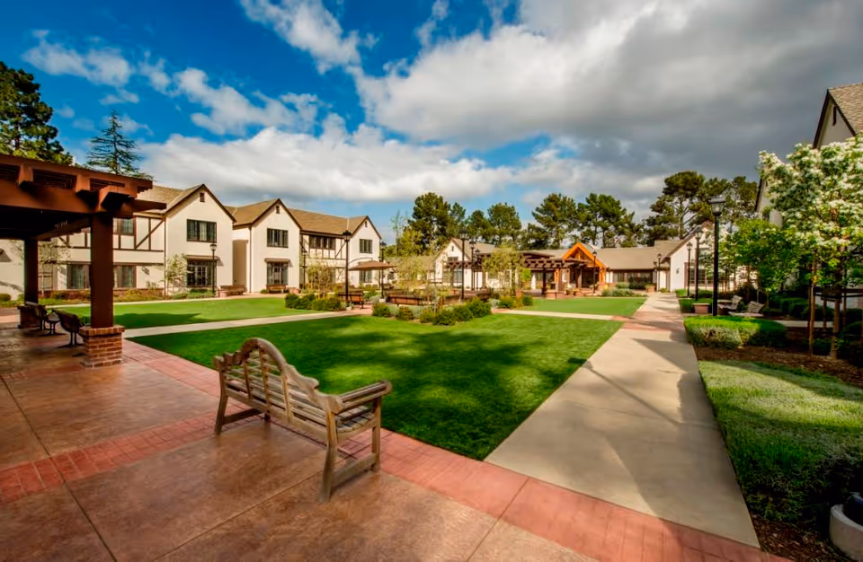 Sunny landscaped courtyard with benches, paved walkways, and surrounding two-story senior living buildings under a partly cloudy sky.