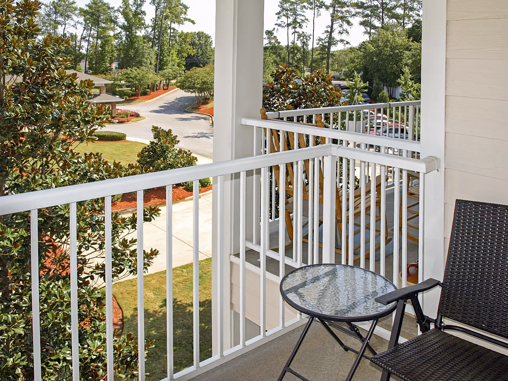 View from a balcony with white railings overlooking a landscaped area with trees, bushes, and a paved driveway. The balcony has a small round glass table and a dark wicker chair, with wooden rocking chairs visible on the adjacent balcony.