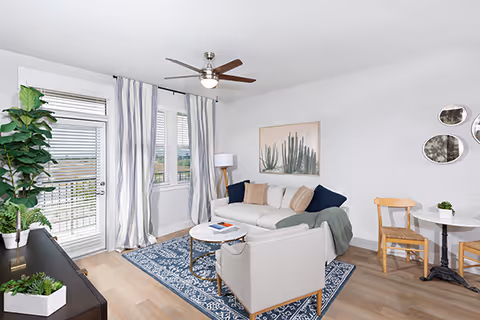 Bright living room with a sofa, armchair, blue patterned rug, small dining table, TV console, potted plant, and sliding glass door to a balcony.