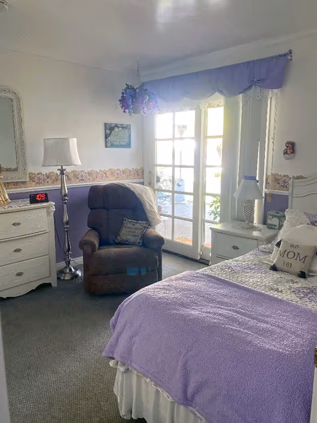 A cozy bedroom with a bed covered in a purple blanket and decorative pillows, including one that says 'Best Mom Ever'. There is a brown recliner chair with a pillow and a light-colored throw blanket. The room has a white dresser with a lamp and a digital clock showing 1:09. French doors with sheer curtains allow natural light to enter the room. The walls are decorated with a floral border and a small framed picture.