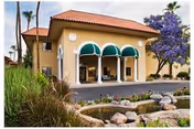 Exterior view of a single-story building with a red tile roof and three green awnings over the entrance. The building is surrounded by landscaping including grass, rocks, and a small pond in the foreground, with palm trees and a purple-flowered tree nearby.