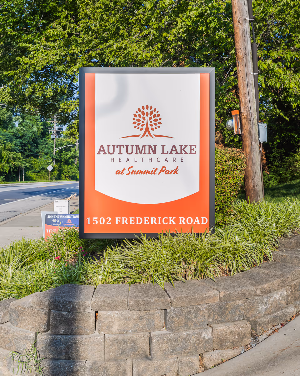 Outdoor sign for Autumn Lake Healthcare at Summit Park located at 1502 Frederick Road, surrounded by greenery and a stone border, with a road and trees in the background.