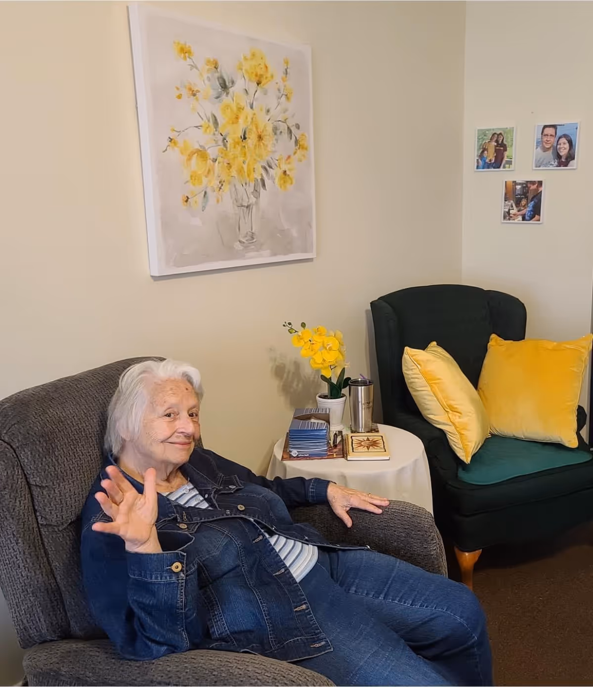 An elderly woman with white hair wearing a denim jacket and jeans is sitting in a gray armchair, waving and smiling. Behind her is a small round table with a yellow orchid plant, books, and a tumbler. To the right, there is a dark green armchair with two yellow pillows. On the wall above the table is a painting of yellow flowers in a vase, and three small family photos are displayed on the adjacent wall.