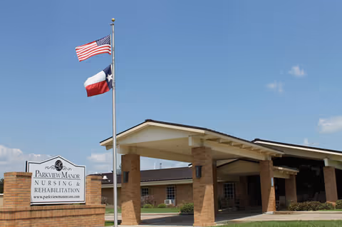 Exterior front view of Parkview Manor Nursing & Rehabilitation building with a covered entrance supported by brick columns. Two flagpoles display the American flag and the Texas state flag. A sign with the facility name and website is visible near the entrance.