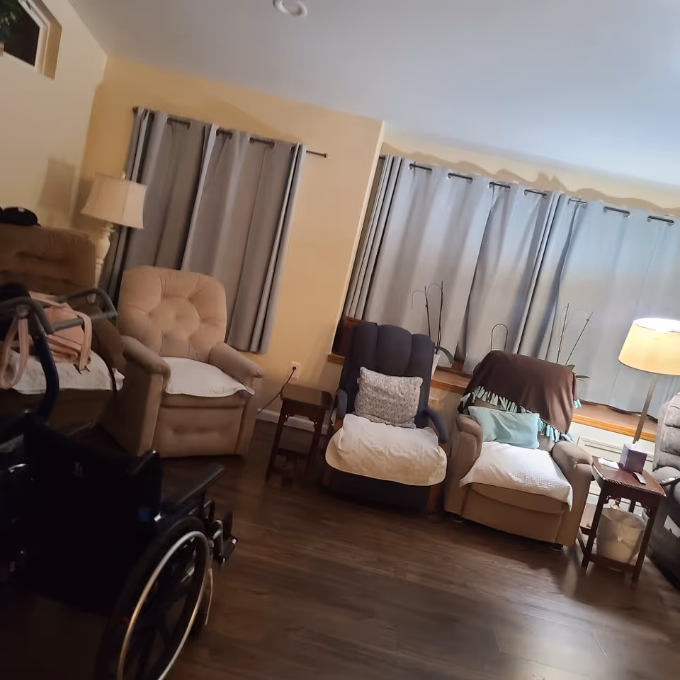 A cozy living room area with several cushioned armchairs, a wheelchair in the foreground, a floor lamp, side tables, and windows with gray curtains. The room has wooden flooring and soft lighting.