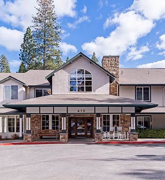 Front exterior view of a senior living facility building with a covered entrance, stone pillars, large windows, and rocking chairs on the porch. Tall pine trees and a partly cloudy sky are visible in the background.