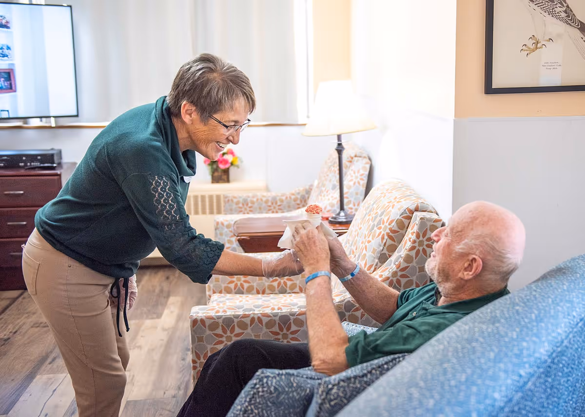 A woman wearing glasses and a green top is smiling and handing a cup with a dessert to an elderly man sitting on a patterned armchair in a cozy living room setting with a lamp and framed artwork on the wall.