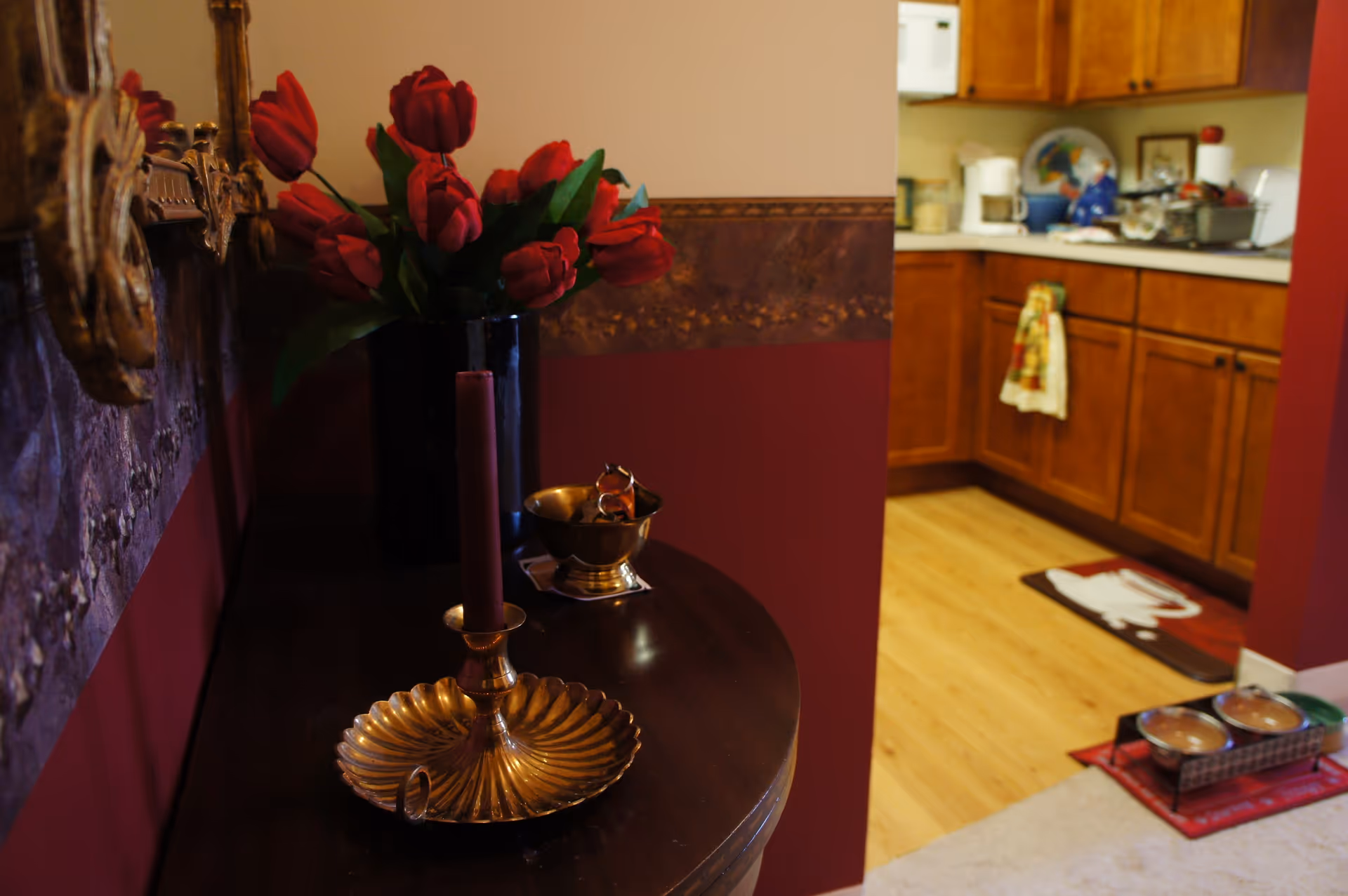 Entry table with red tulips, a brass candle holder and bowl in the foreground and a kitchen with wooden cabinets visible in the background.