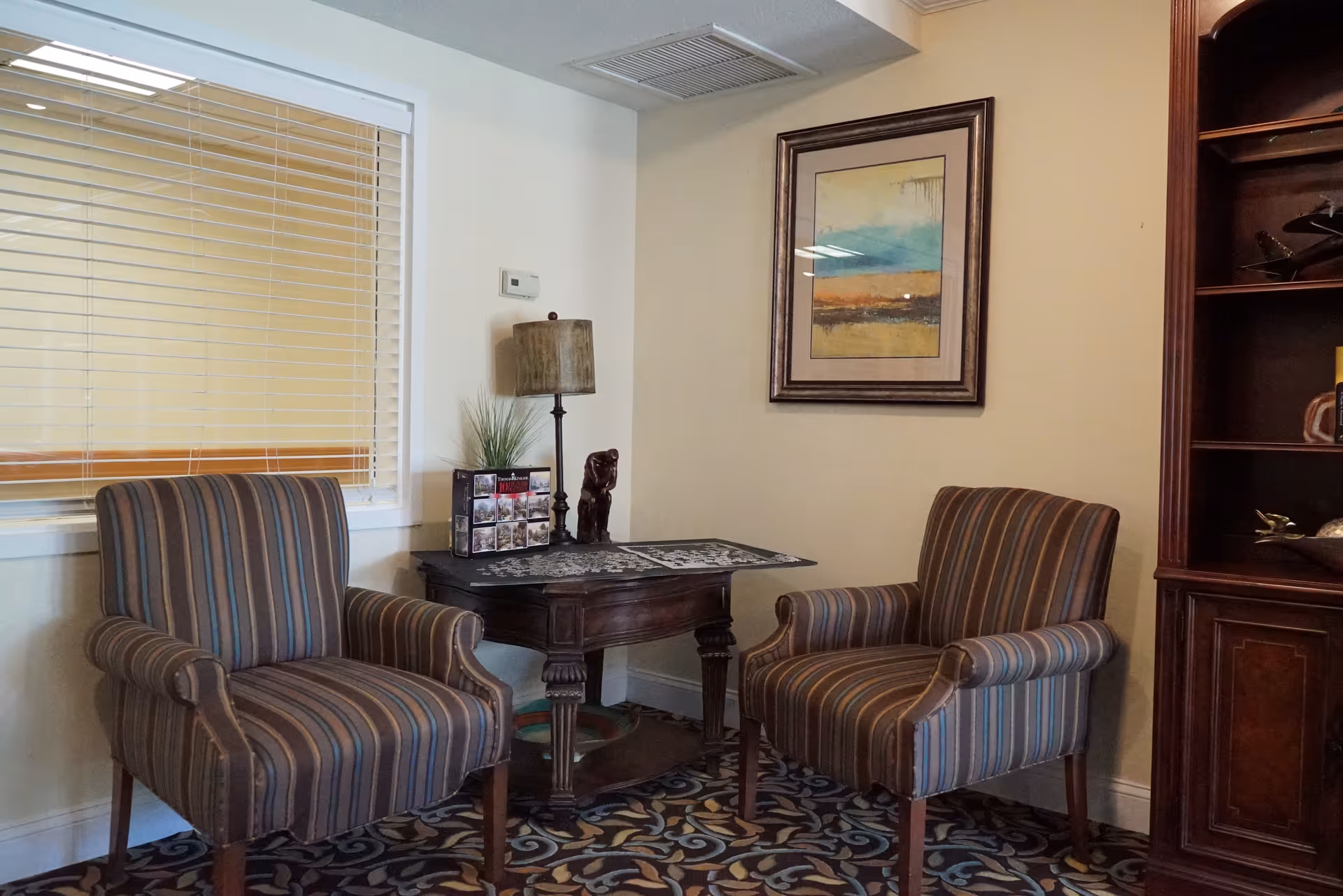 A cozy sitting area with two striped armchairs facing each other, separated by a dark wooden table with a lamp, a small plant, and decorative items. A framed abstract painting hangs on the beige wall above one chair, and a wooden bookshelf is partially visible on the right side. The floor is covered with a patterned carpet, and a window with closed blinds is on the left wall.