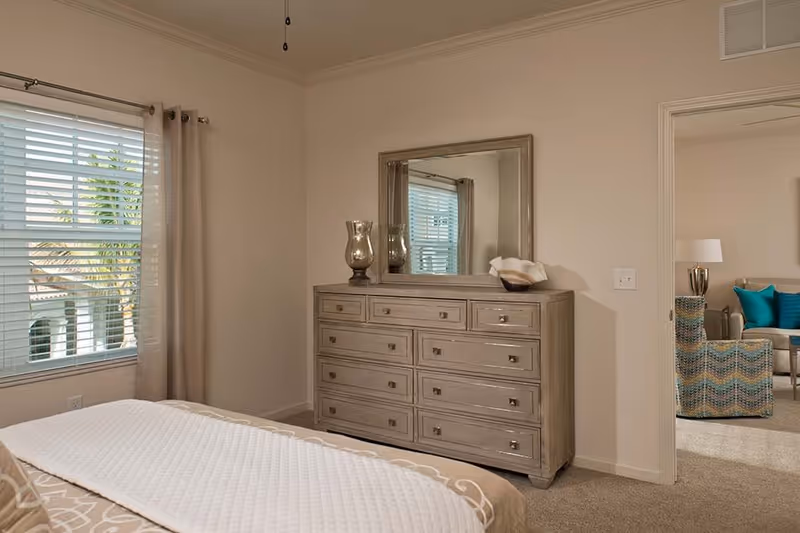 A bedroom with a bed partially visible in the foreground, a large wooden dresser with multiple drawers and a mirror above it against the wall, a window with blinds and beige curtains letting in natural light, and a doorway leading to a living room area with a chair, sofa, and lamp.