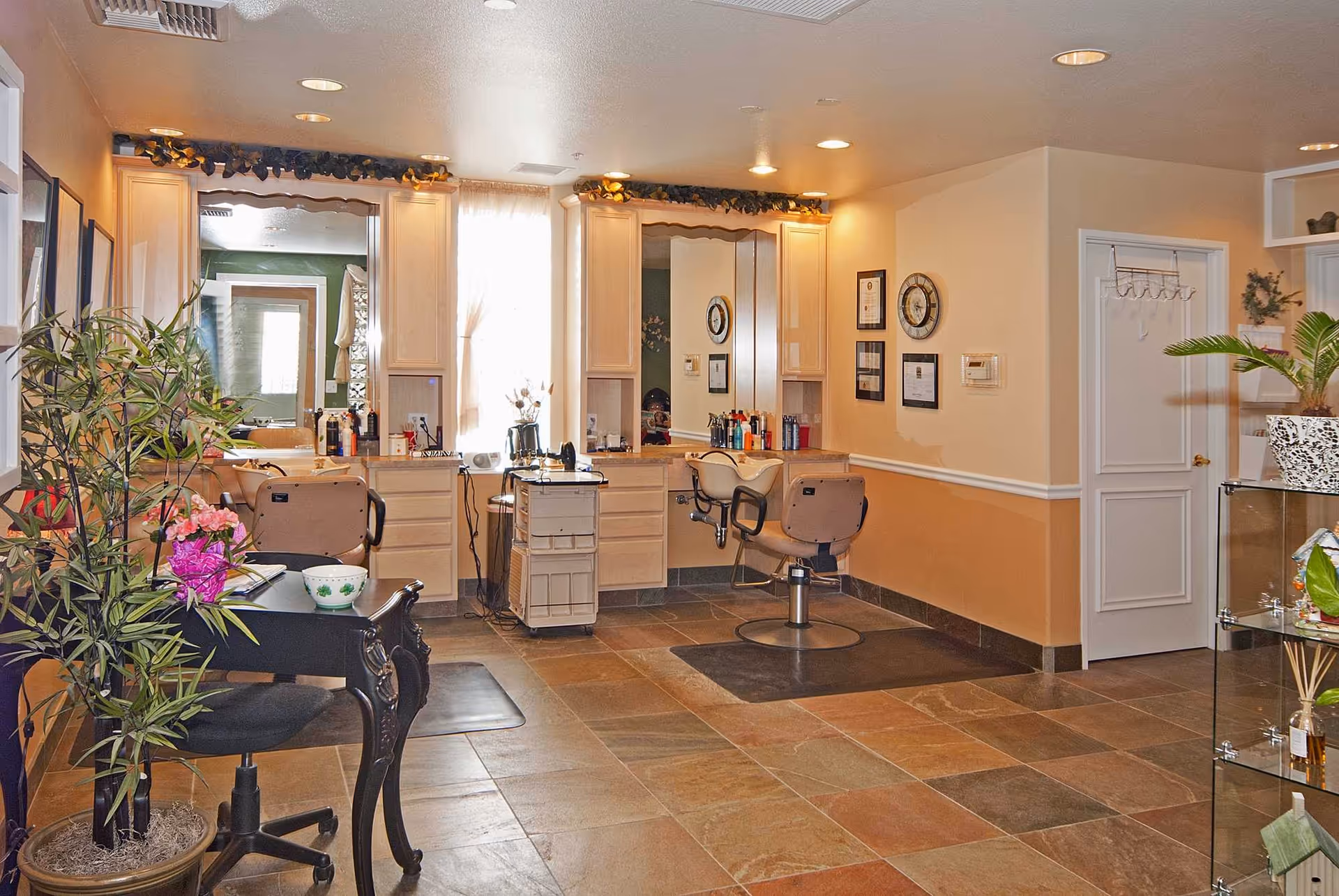 Well-lit salon area with styling chairs, mirrors, countertop stations, plants and a tiled floor.