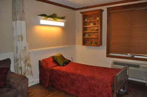 A small bedroom in a senior care facility with a red patterned bedspread and wooden furnishings.