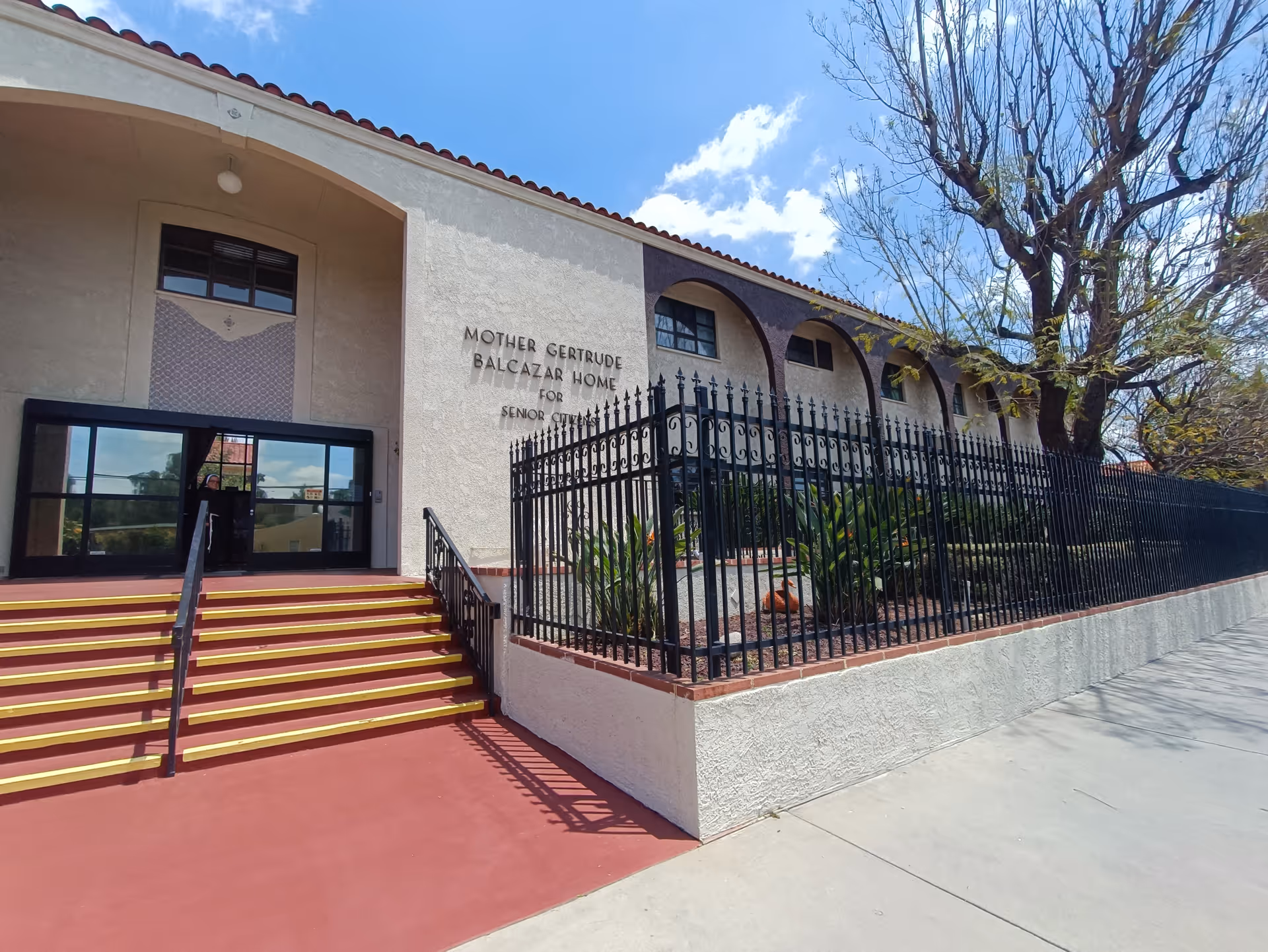 Front entrance of the Mother Gertrude Balcazar Home showing red steps, an iron fence, and signage on a two-story stucco building under a blue sky.