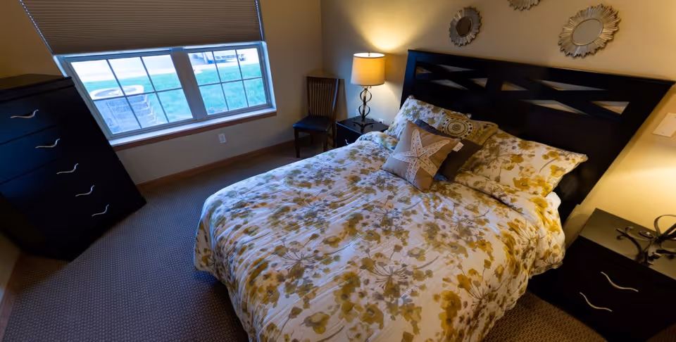 Bedroom with a floral-patterned bed, dark wood headboard and matching dressers, a nightstand lamp, and a window.