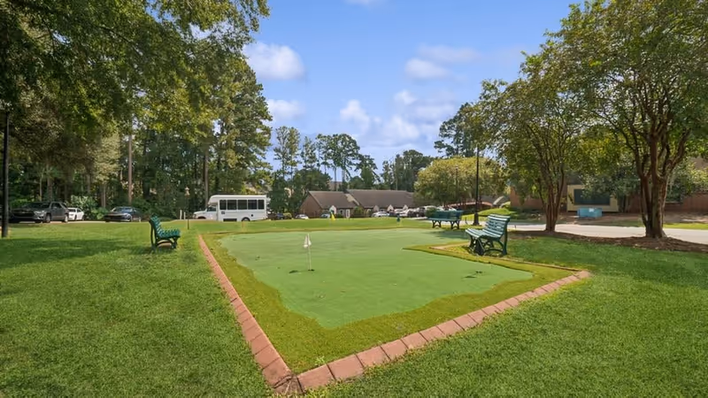 A small outdoor putting green with benches, trees, parked vehicles and residential buildings in the background.