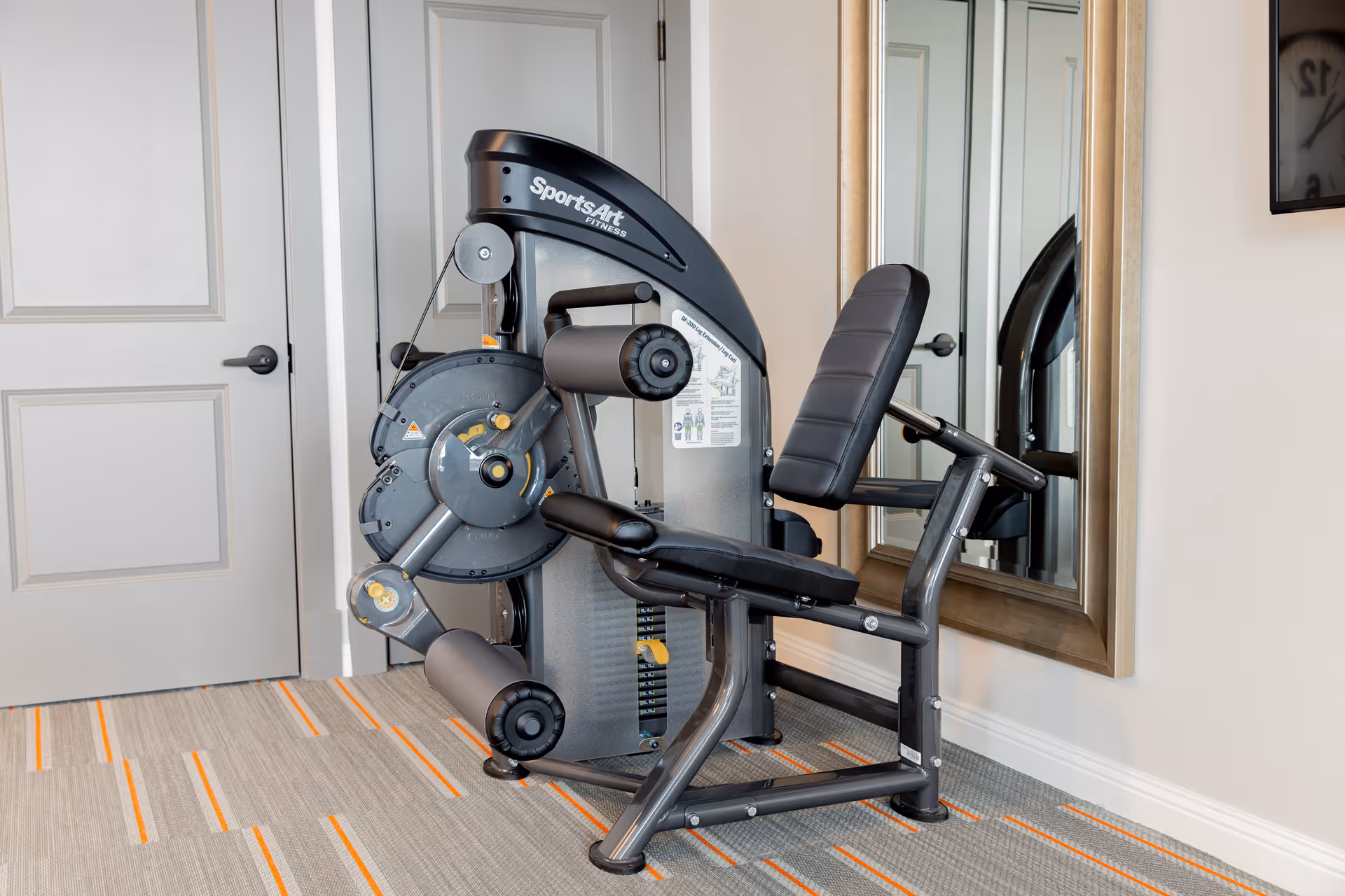 A leg extension exercise machine in a small fitness room next to a mirror and a closed door.