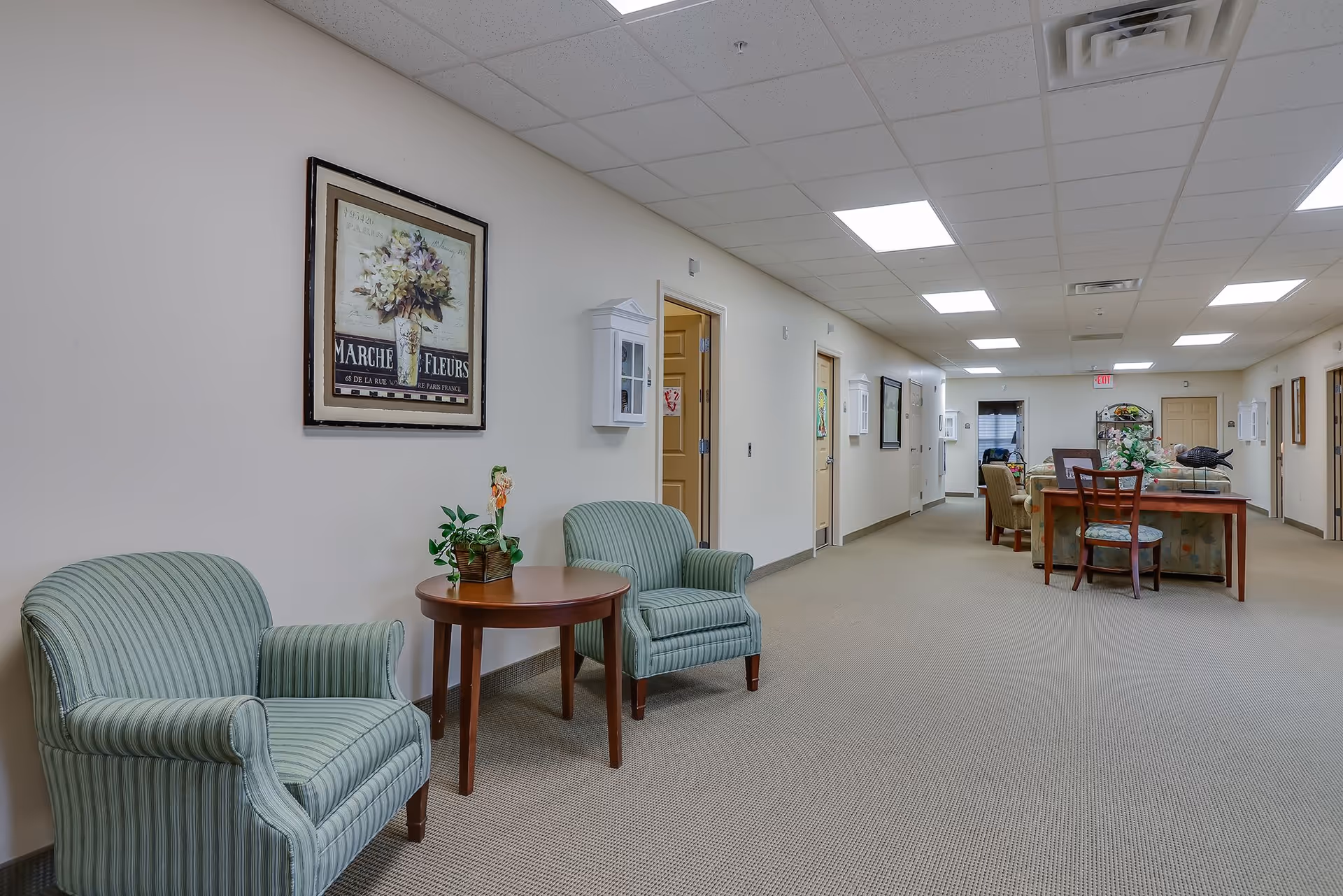 Bright hallway common area with upholstered chairs and a small table in the foreground and a dining table further down the corridor.