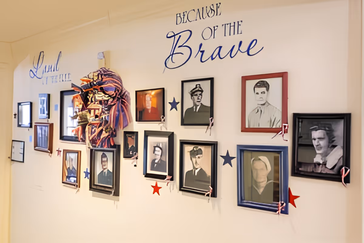 Interior hallway wall display with framed portraits, patriotic star decorations and a colorful fabric mask beneath the words "Because of the Brave".