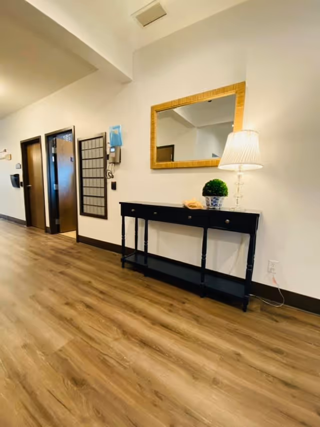 A hallway in a senior living facility with wooden flooring, white walls, and dark brown baseboards. On the right side, there is a black console table with three drawers, a small green potted plant, a decorative item, and a table lamp with a white shade. Above the table is a rectangular mirror with a golden frame. On the left side of the hallway, there are two open doors and a wall-mounted telephone with a blue cover.