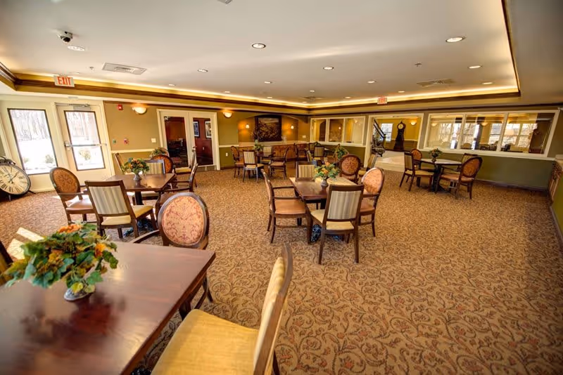 A spacious dining room with multiple wooden tables and chairs arranged neatly on a patterned carpet. The room is well-lit with ceiling lights and natural light coming through large windows and glass doors. There are floral centerpieces on the tables and a large clock leaning against the wall near the exit door.