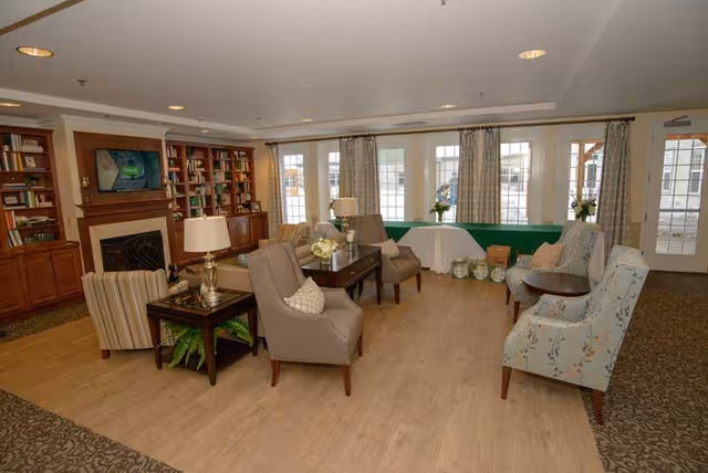A cozy living room area in North Woods Village at Edison Lakes featuring multiple upholstered armchairs arranged around wooden side tables and a central coffee table with flower arrangements. The room has large windows with curtains, built-in wooden bookshelves, a fireplace with a mounted TV above it, and a light wood floor with carpeted edges.