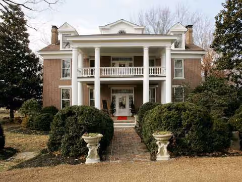 Brick two-story manor exterior with columns, a central porch, and manicured hedges.