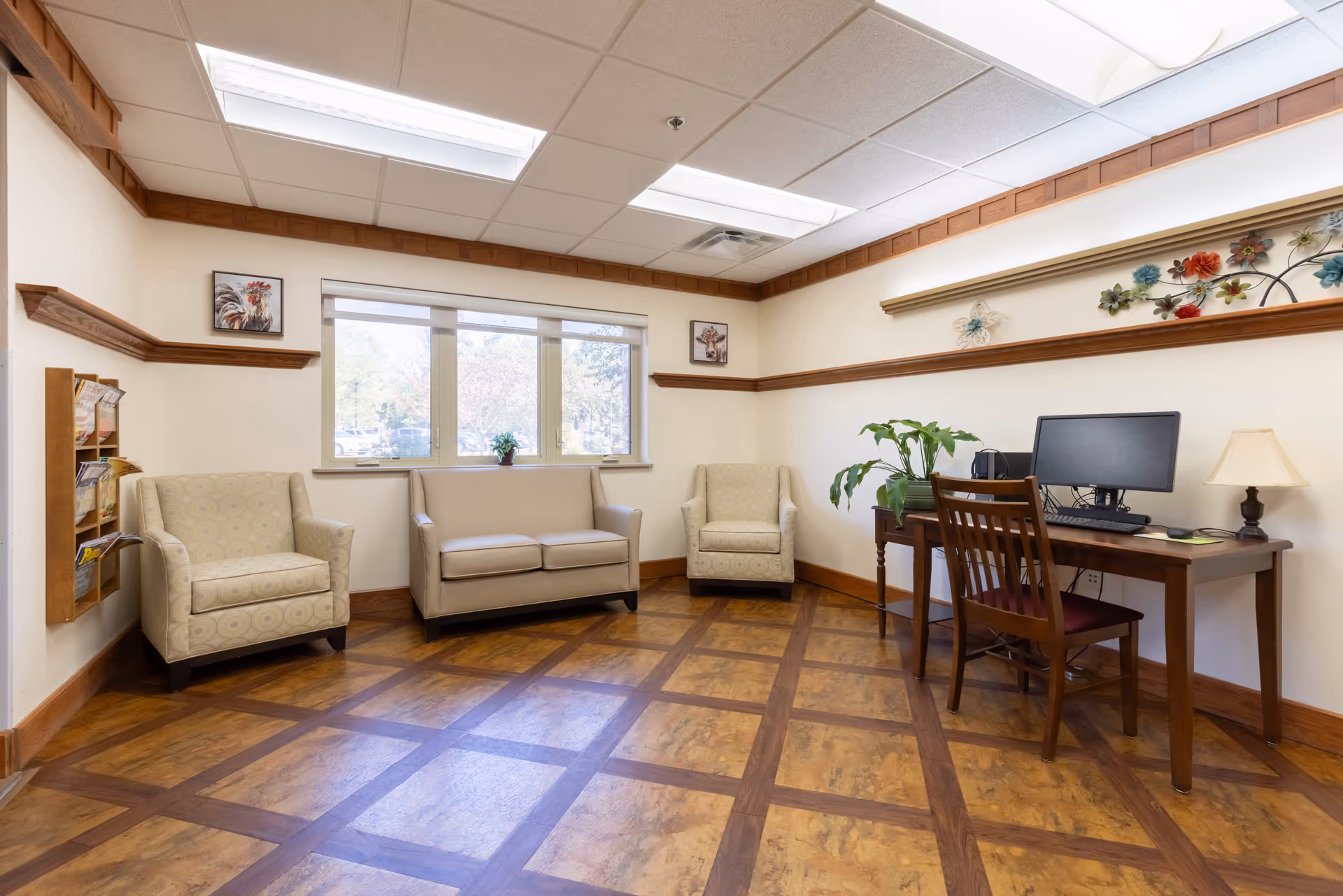 A bright and clean common area with two beige armchairs and a beige loveseat arranged near a window. A wooden desk with a computer, lamp, and plant is positioned against the wall. The room features wood trim, patterned wood flooring, and decorative wall art including a floral metal piece and framed pictures.