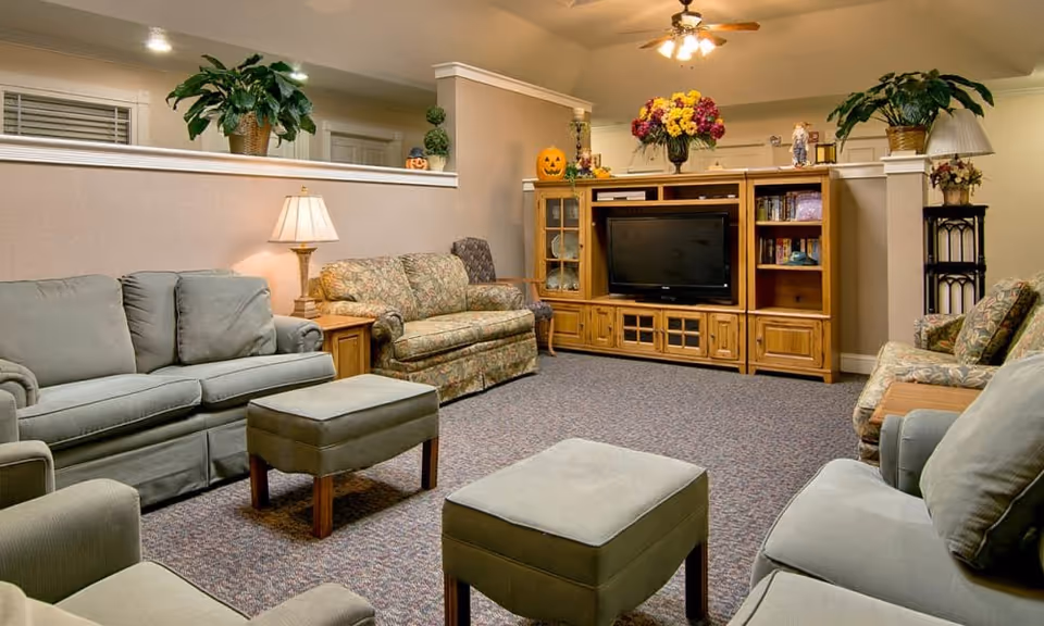 A cozy senior living facility common area with multiple upholstered sofas and armchairs arranged around two ottomans. A wooden entertainment center with a TV, books, and decorative items is against the wall. The room is decorated with plants, flowers, and Halloween-themed pumpkins, and is softly lit by a table lamp and ceiling fan lights.