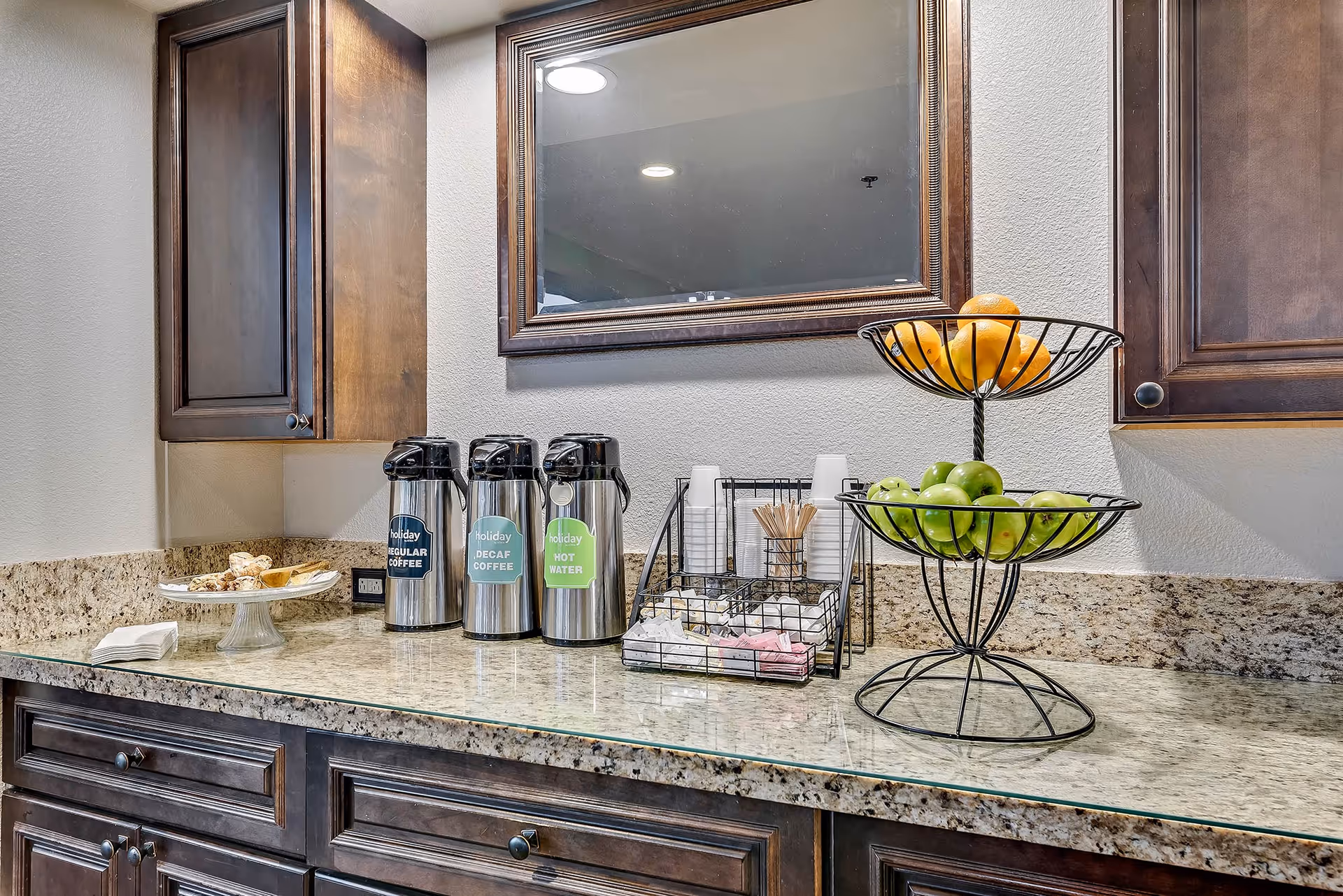 A kitchenette countertop with three coffee dispensers, disposable cups and condiments, and a two-tier fruit basket beneath a wall mirror.
