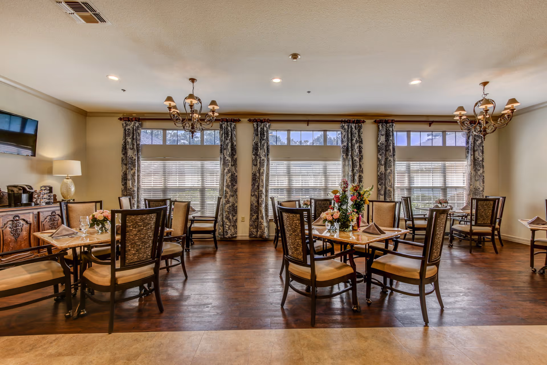 A senior living facility dining room with several tables and chairs arranged neatly. Each table has a floral centerpiece and folded napkins. Large windows with patterned curtains let in natural light, and two chandeliers hang from the ceiling. A sideboard with a lamp and coffee station is visible on the left side of the room.