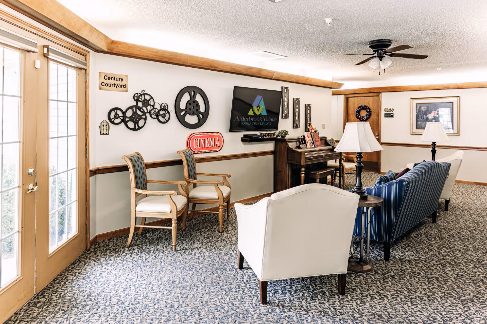 A cozy assisted living common area with a piano, several chairs, a striped couch, and two white armchairs arranged around side tables with lamps. The wall features decorative film reels, a 'Cinema' sign, and a flat screen displaying the Alderbrook Village Assisted Living logo. A door with glass panels leads outside, and the room has carpeted flooring and wood trim accents.