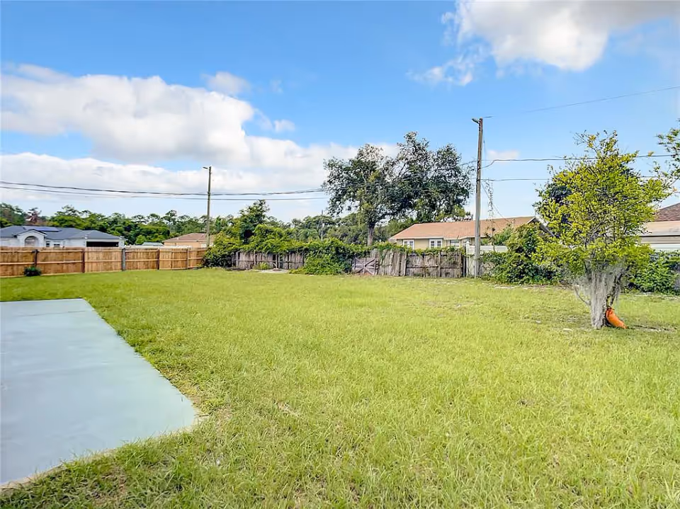 A large grassy backyard area with a small tree on the right side and a concrete patio on the left. The yard is enclosed by wooden fences, with houses and trees visible in the background under a partly cloudy blue sky.