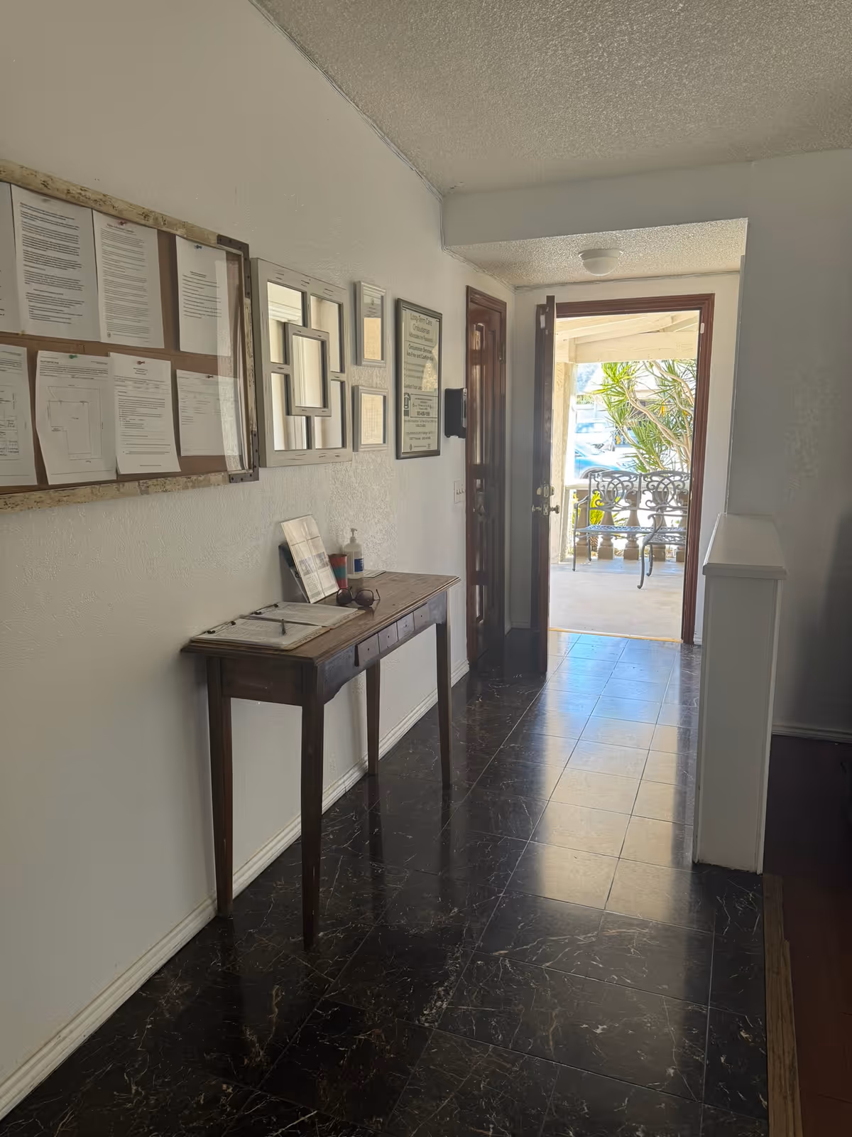 Narrow interior hallway with a small wooden table, wall bulletin board and an open front door leading to an outdoor seating area.