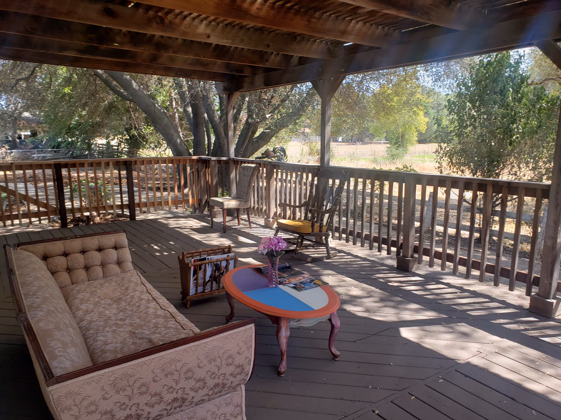 Covered wooden deck with a sofa, chairs and a small colorful table overlooking trees and a yard.