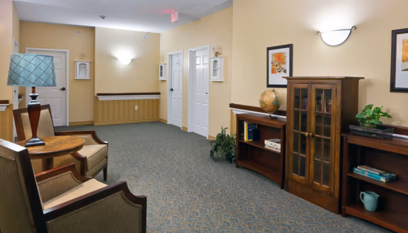 A carpeted interior common area with armchairs and a side table with a lamp, wooden bookshelves and cabinets against beige walls.