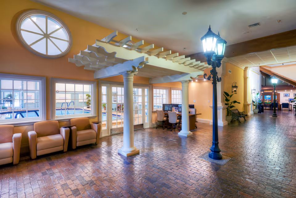 Interior view of a senior living facility featuring a spacious hallway with brick flooring, beige walls, and a decorative white pergola supported by columns. There are three beige armchairs near windows that look into an indoor pool area. A desk with computer monitors and chairs is situated under the pergola. The hallway is illuminated by vintage-style street lamps and has benches and plants along the walls.