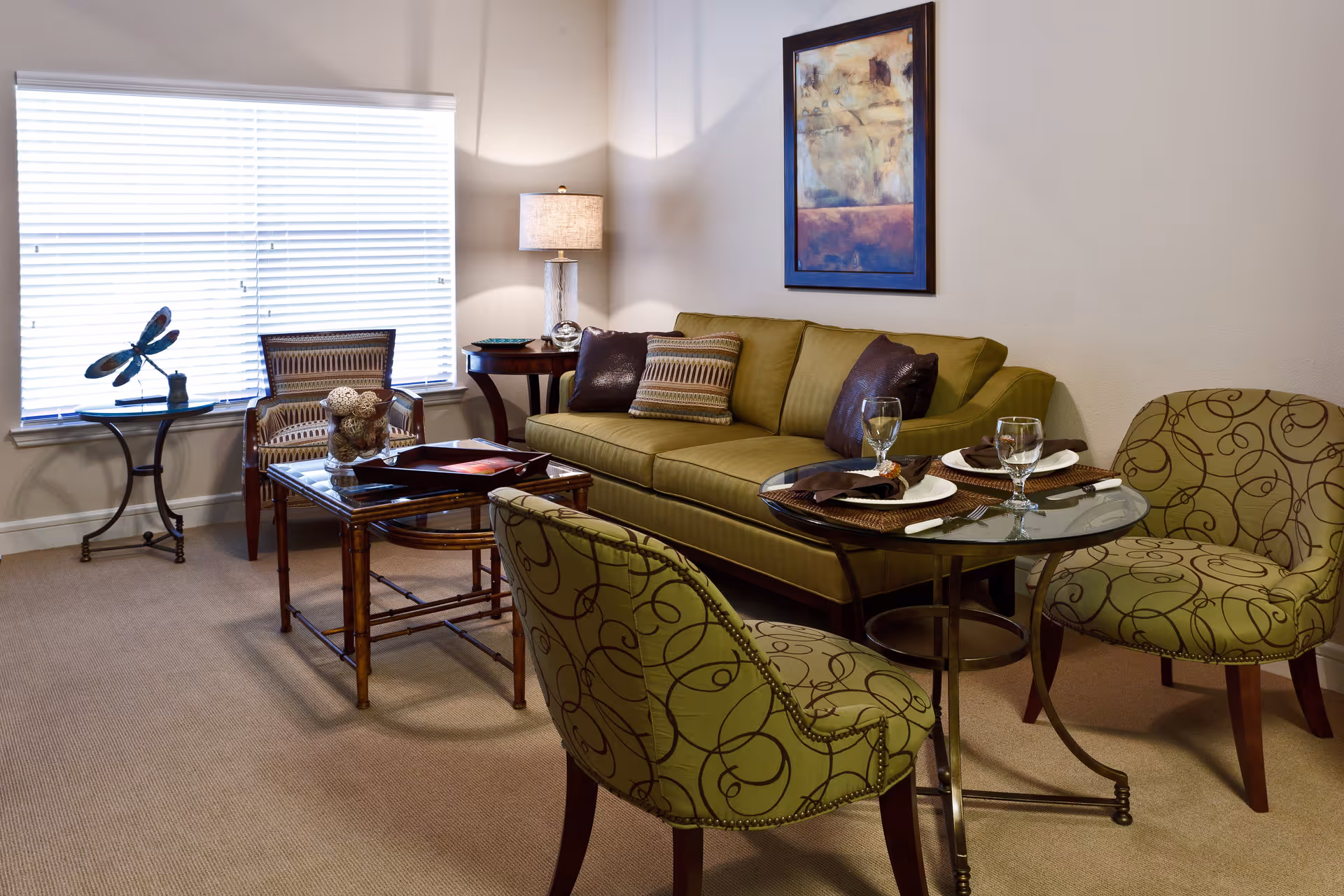 A cozy living room with a green sofa adorned with pillows, two patterned green chairs around a small glass dining table set with plates, napkins, and glasses. There is a wooden coffee table with decorative items, a side table with a lamp, and a large window with blinds letting in natural light. A framed abstract painting hangs on the wall above the sofa.