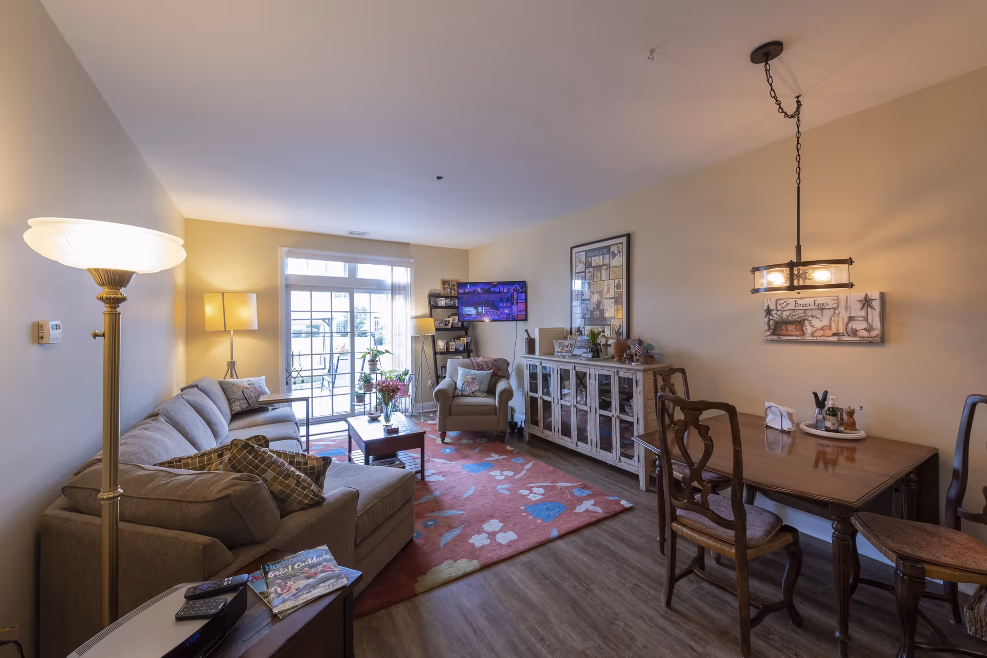 A cozy living room with a gray sectional sofa, a wooden coffee table on a red floral rug, an armchair, and a wooden dining table with chairs. The room has warm lighting from floor and table lamps, a wall-mounted TV, a glass-front cabinet with decorative items, and a large window with a door leading outside.