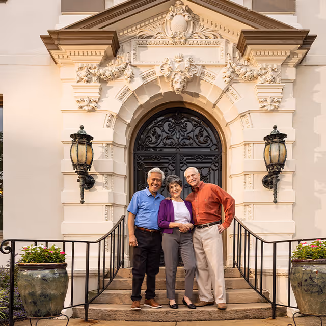 Three older adults smiling and standing on the front steps of an ornate mansion entrance with a decorative arched doorway.