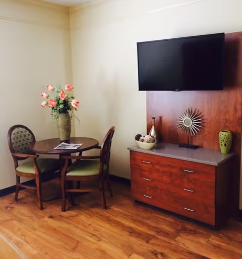 A small seating area with a round wooden table and three chairs with green cushions. A vase with pink flowers is on the table. To the right, there is a wooden cabinet with three drawers and decorative items on top, including a sunburst sculpture and a green vase. A flat-screen TV is mounted on the wall above the cabinet.