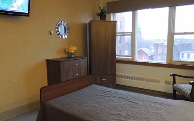 A simple bedroom with a bed covered in a gray blanket, a wooden dresser with a small vase of yellow flowers, a tall wooden cabinet, a chair with striped upholstery, and a wall-mounted TV. Large windows show a view of buildings and a church steeple outside.