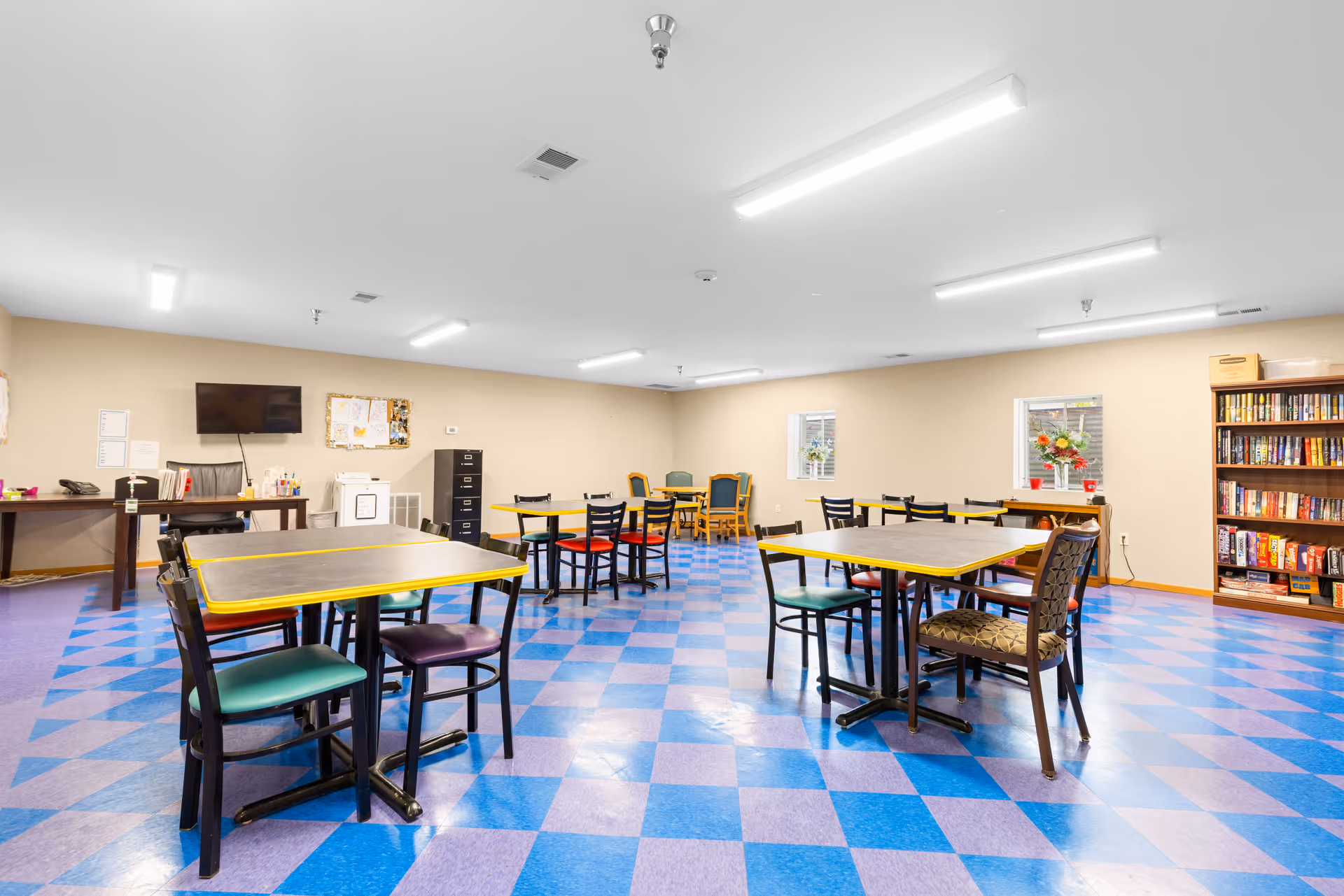 A spacious room with multiple tables and chairs arranged for group seating. The floor has a blue and purple checkered pattern. There is a bookshelf filled with books and board games on the right side, and a desk with a computer and office supplies on the left. The walls are light beige with two small windows, and the ceiling has several fluorescent lights.