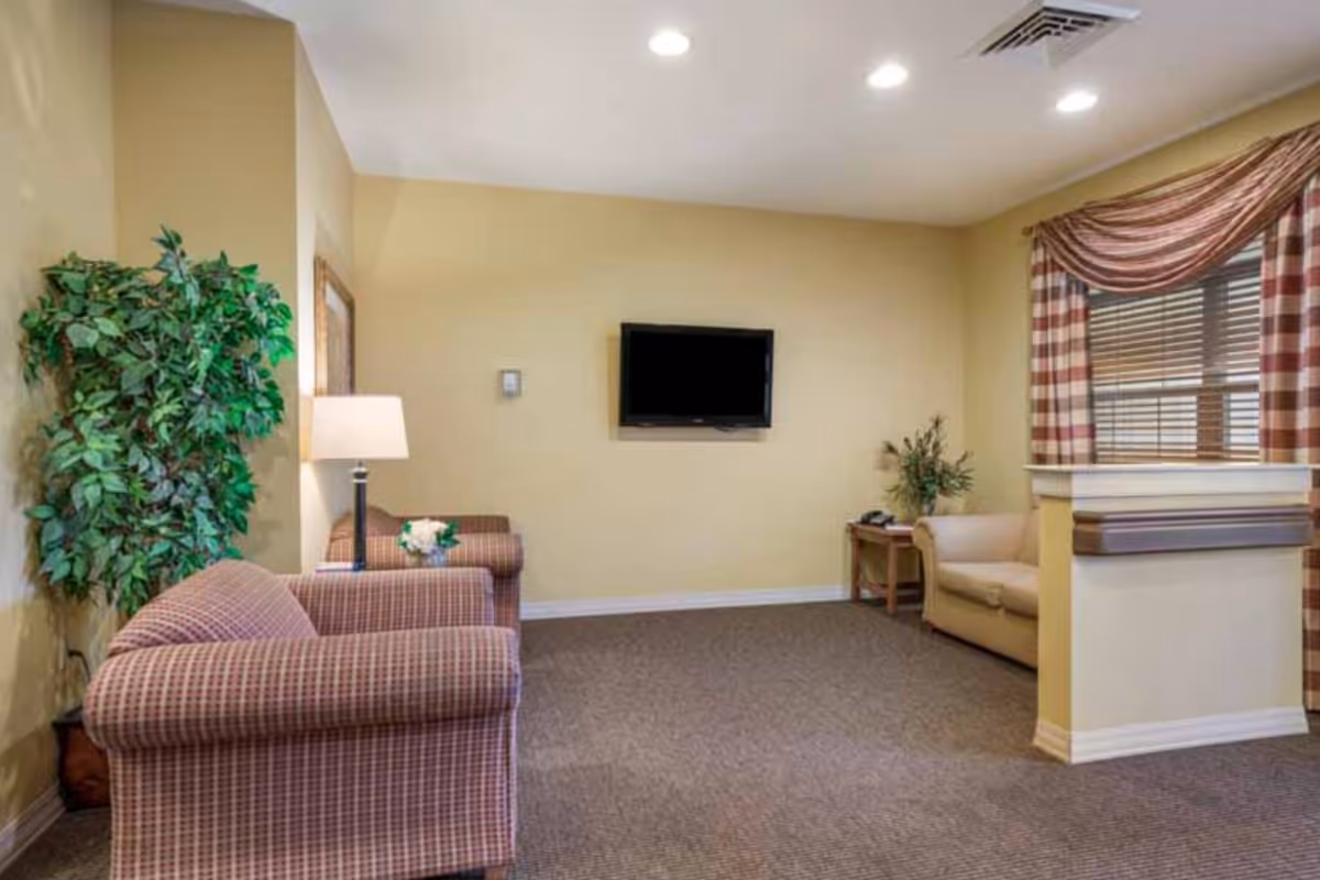 A cozy living room area with beige walls and carpeted floor. The room features two patterned armchairs on the left side, a beige loveseat on the right, a small wooden side table with a telephone and plant, a floor lamp, a large potted plant, and a flat-screen TV mounted on the wall. A window with red and white checkered curtains is visible on the right side.